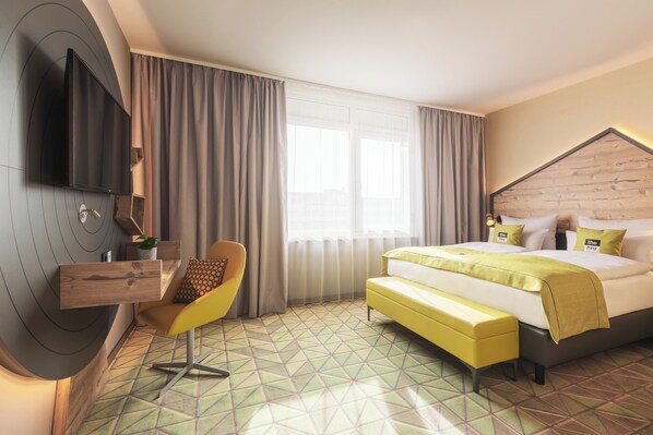 A modern hotel room with a large bed, a TV mounted on the wall, a yellow chair, and a geometric patterned floor.