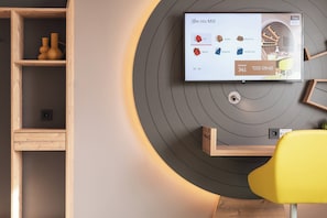 A modern living room with a TV mounted on a circular wall, wooden shelves, and a yellow chair. - Holiday Inn – the niu, Mill Cologne Mülheim by IHG (Cologne)