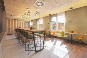 A modern dining area with a long wooden table and multiple bar stools, large windows providing natural light, and a ceiling with a unique geometric light fixture. - Holiday Inn – the niu, Mill Cologne Mülheim by IHG (Cologne)