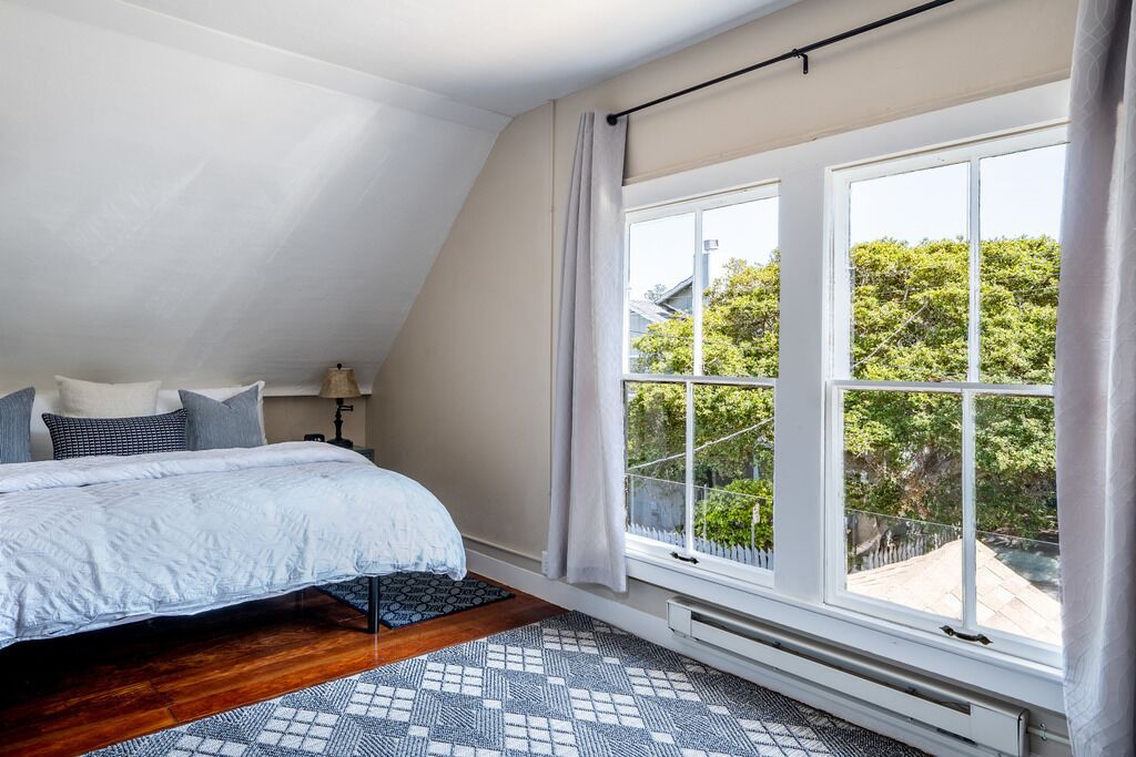 A bedroom with a sloped ceiling, a bed with a white duvet, a patterned rug, and a large window with a view of greenery outside.