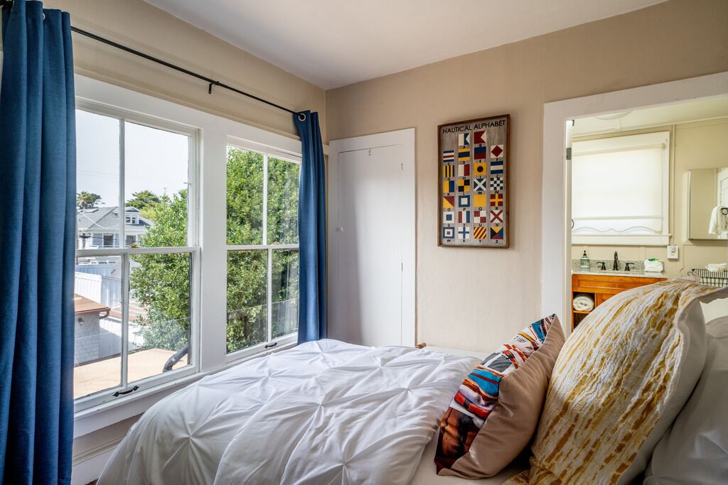 A bedroom with a large bed, a window with curtains, a nautical alphabet wall art, and a bathroom visible through an open door.