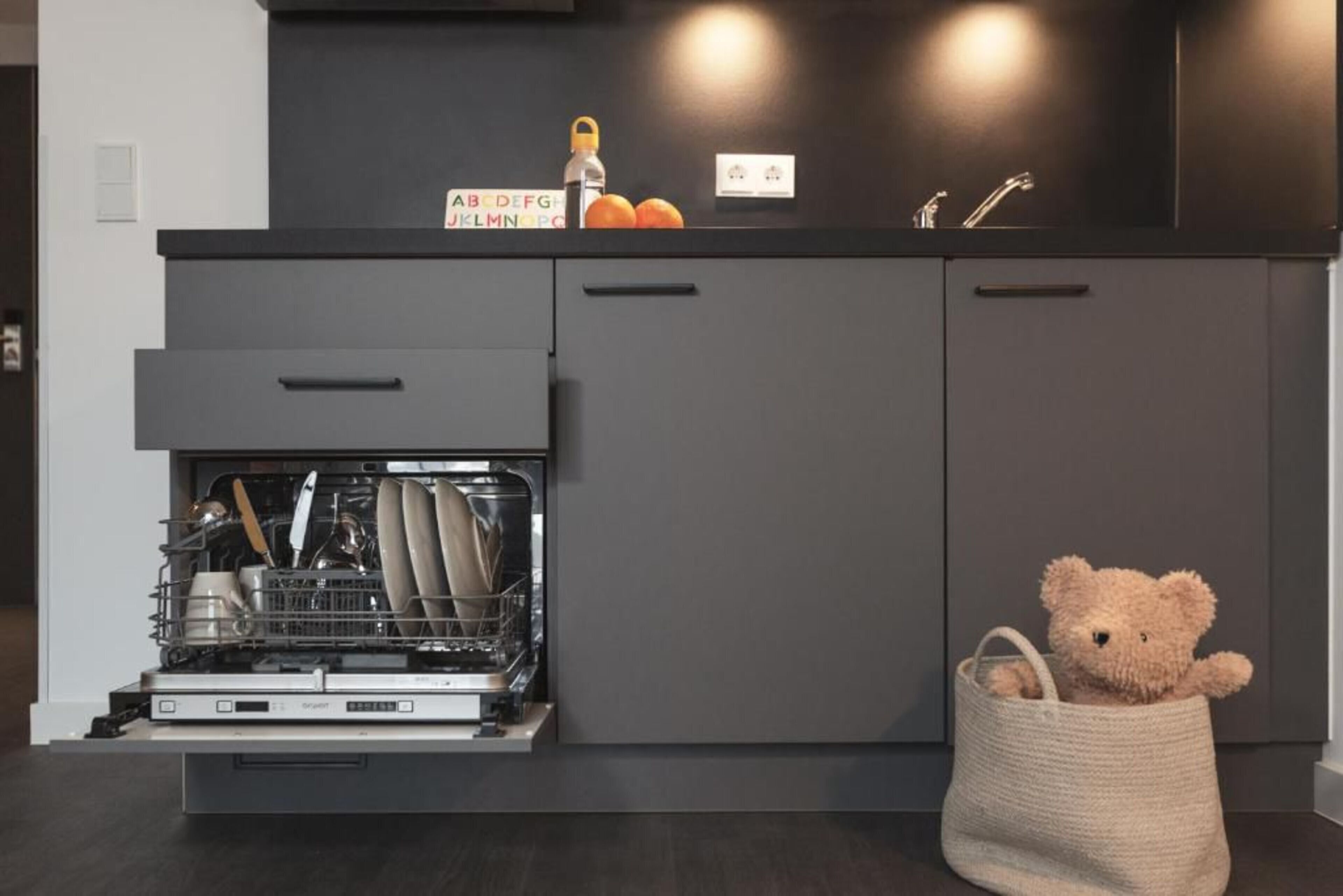 Una cocina moderna con armarios grises, lavavajillas y un osito de peluche en una cesta.