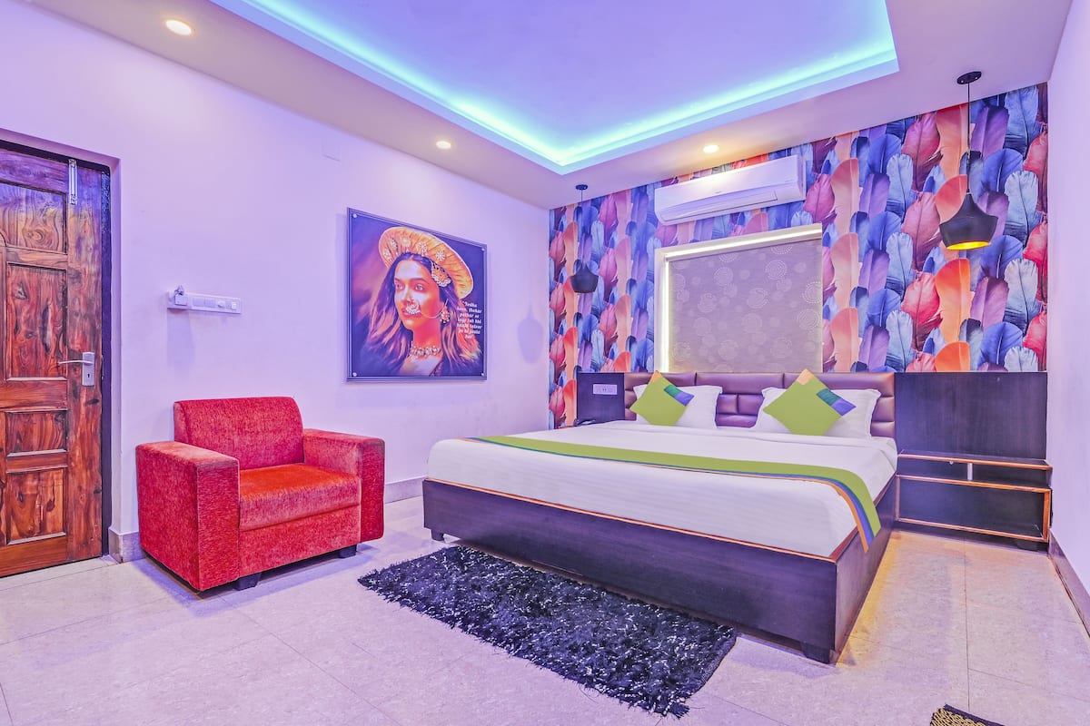 A modern bedroom with a wooden door, a red chair, a bed with green and white bedding, and a colorful feather-patterned wall.