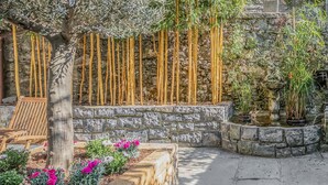 Un muro de piedra con plantas de bambú, un banco de madera y un cantero con flores rosas.