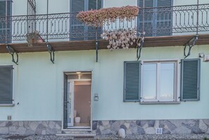 Un edificio con base de piedra, un balcón de madera con barandilla metálica y una puerta que da a un espacio interior.