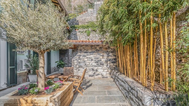 Una escalera de piedra conduce a una terraza de madera con mobiliario exterior, rodeada de plantas de bambú y un muro de piedra.