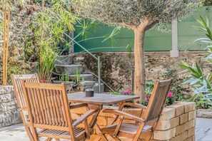Un patio al aire libre con mesa y sillas de madera, rodeado de plantas y un muro de piedra.