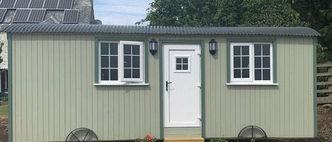 A small, single-story building with a white door and two windows, each with a white frame. There are two lantern-style lights on either side of the door. The building is painted in a light green color and has a gravel driveway.