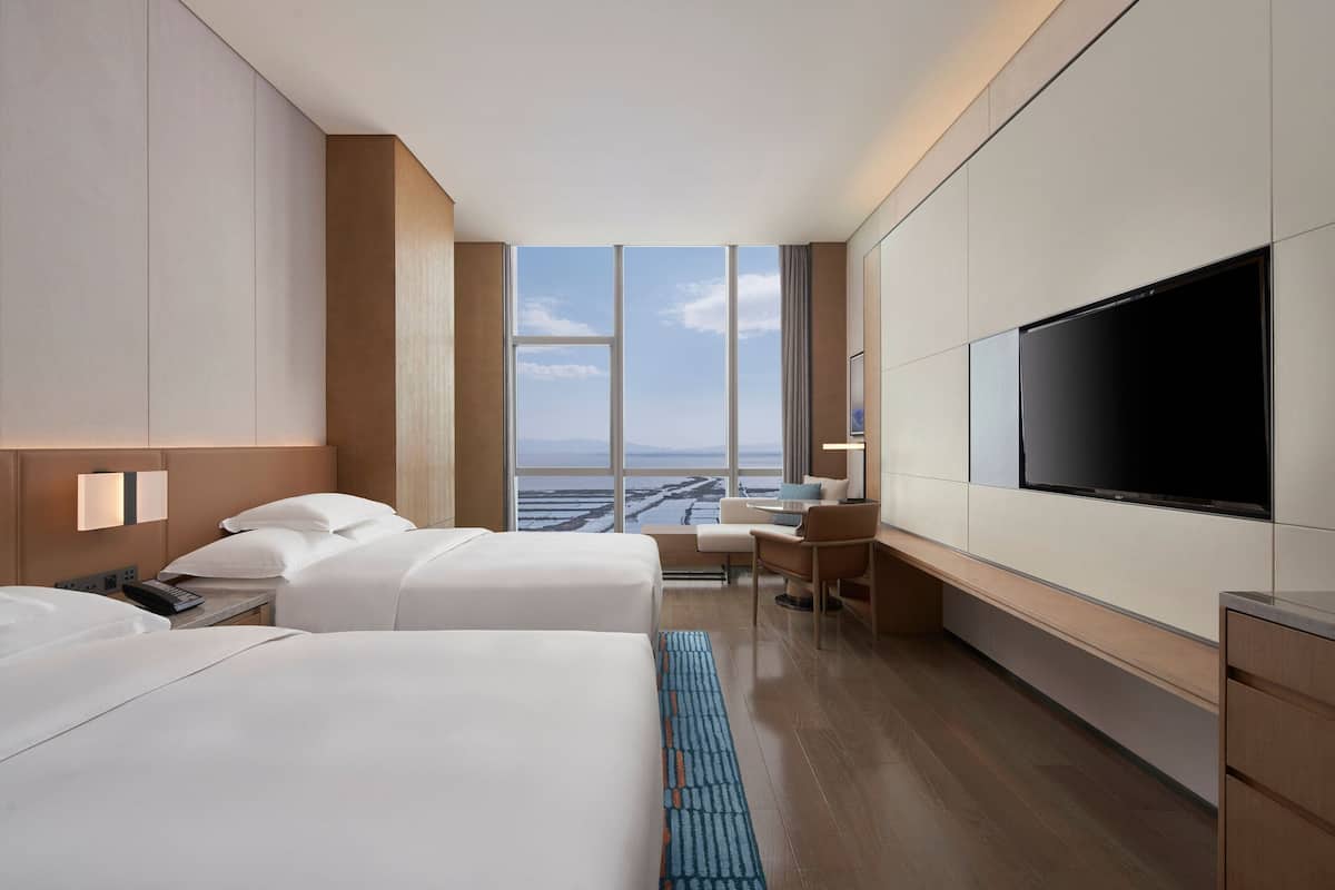 Quarto de hotel moderno com duas camas, uma escrivaninha, televisão e uma janela grande com vista para a cidade.