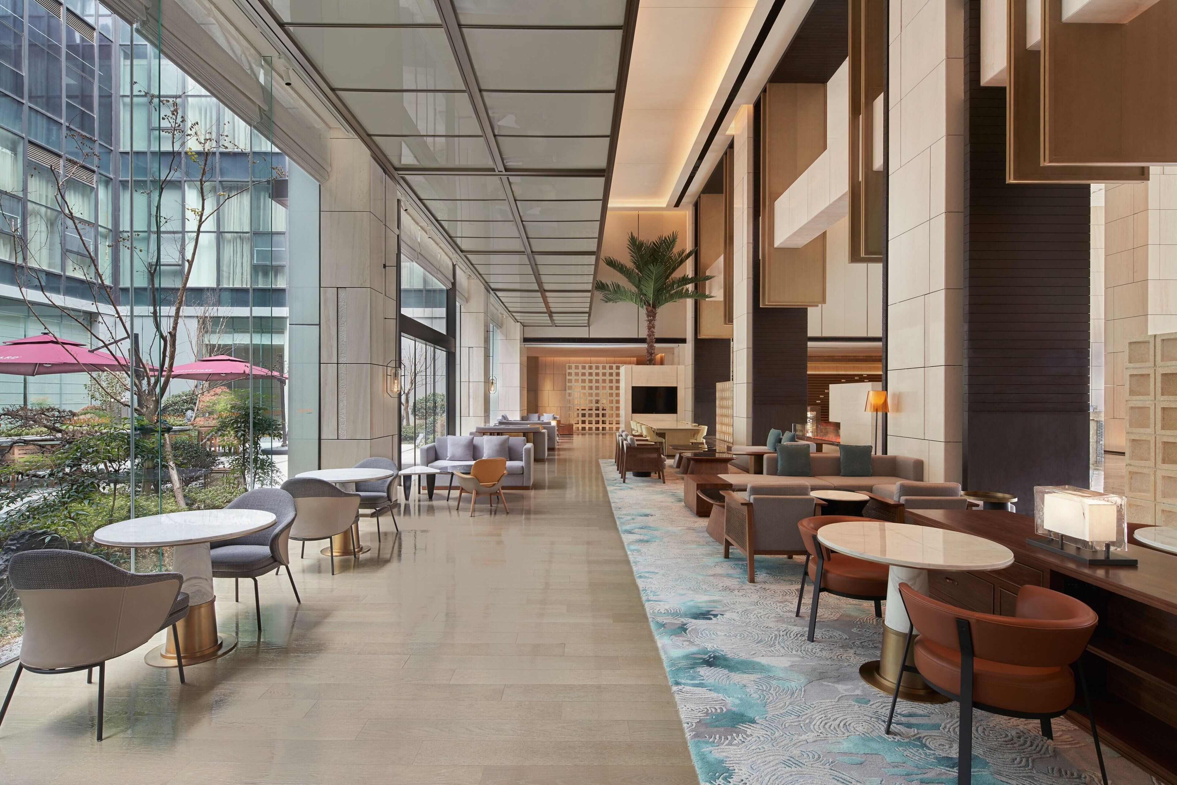 Un hall d’hôtel moderne avec des tables et des chaises rondes, une grande fenêtre offrant une vue sur la ville et un tapis à motifs.