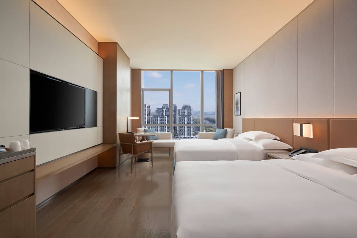 Quarto de hotel com duas camas, televisão de tela plana, escrivaninha e uma grande janela com vista para a cidade.
