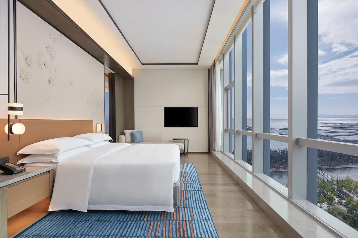 Quarto de hotel com uma cama grande, televisão de tela plana, mesa de cabeceira com telefone e vista para a paisagem urbana através de grandes janelas.