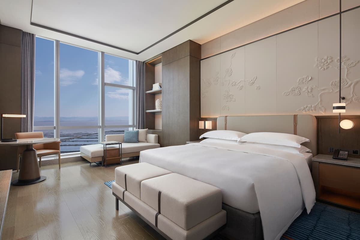 Quarto de hotel moderno com uma cama grande, área de estar com vista para o oceano e escrivaninha com abajur.
