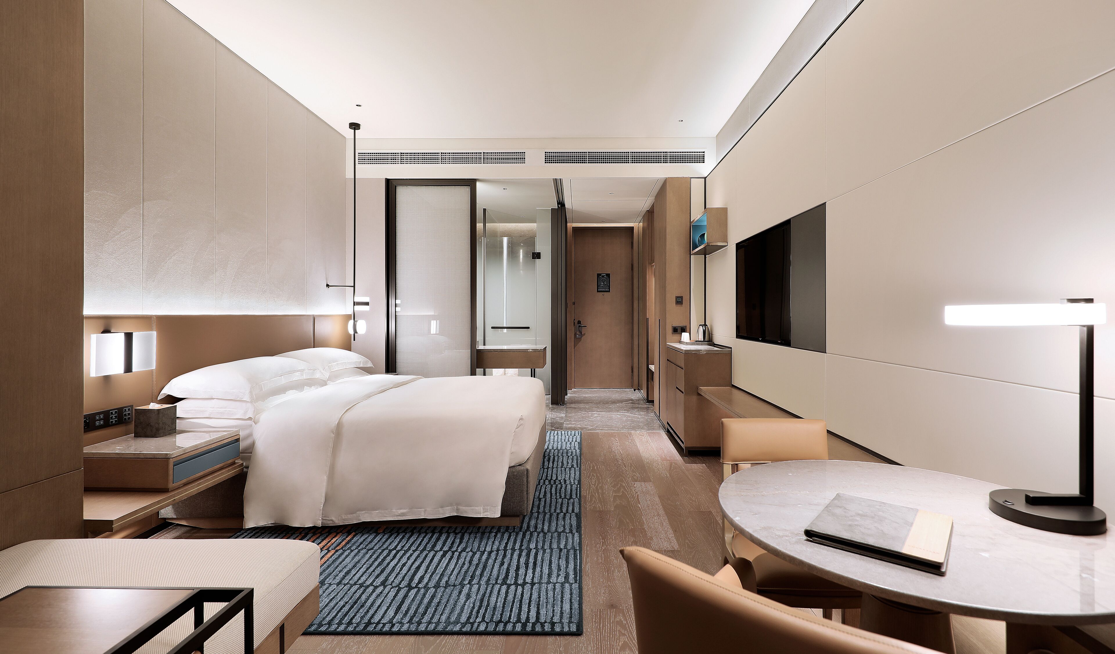Quarto de hotel moderno com cama, mesas de cabeceira, escrivaninha, poltrona, TV e banheiro visível por uma porta aberta.