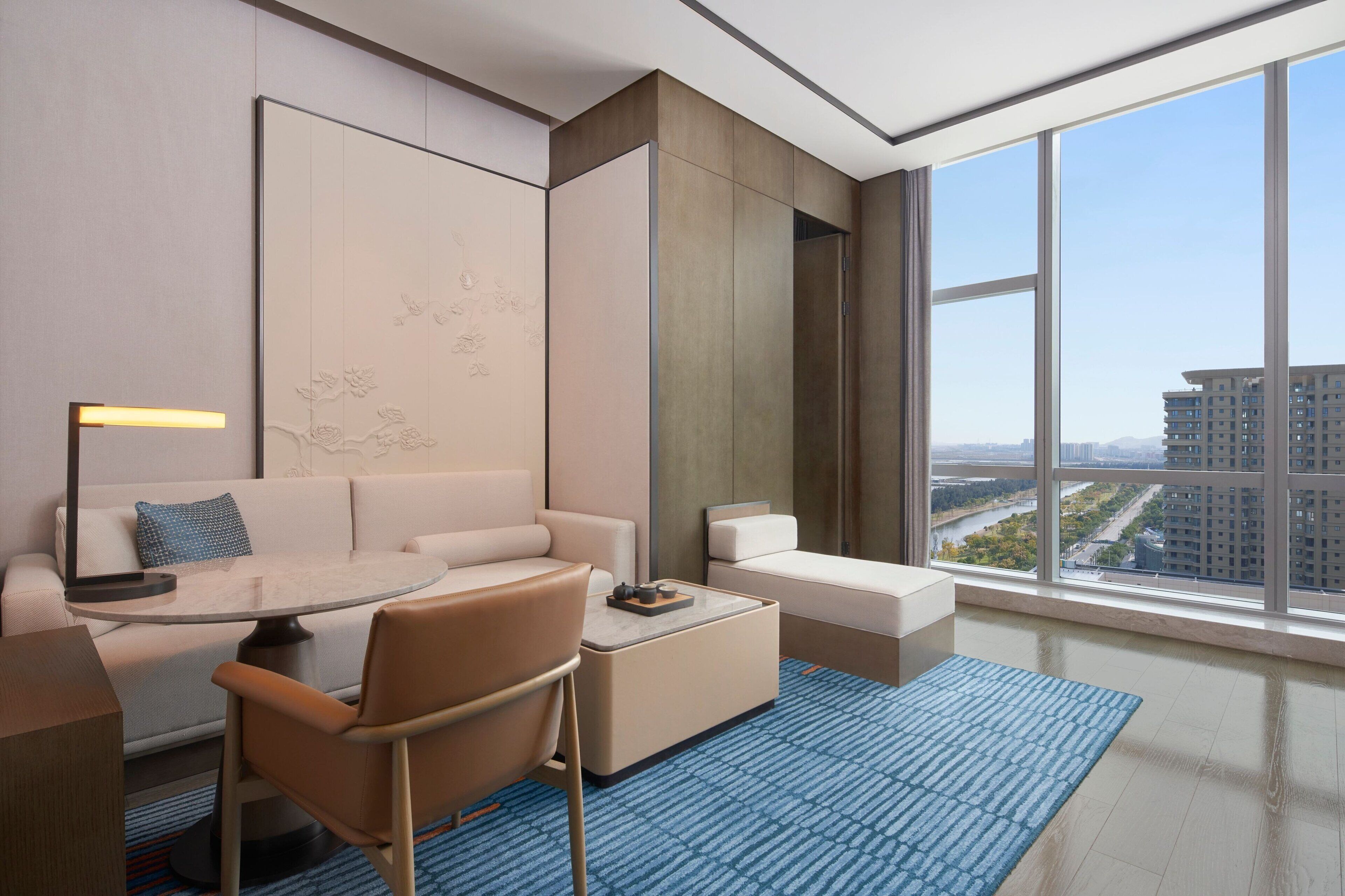 A modern hotel room with a large window, a round table, a sofa, and a coffee table.