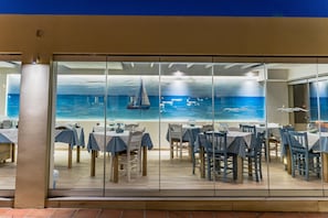 A restaurant with a view of the ocean through large glass windows, tables with blue tablecloths, and a sailboat in the background.
