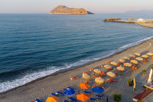 A beach with rows of sun loungers and umbrellas, a rocky outcrop in the water, and a distant view of a city.