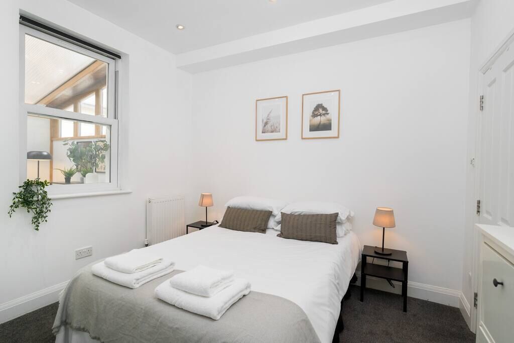 A neatly arranged bedroom with a bed, two bedside tables with lamps, a window with a plant, and two framed pictures on the wall.