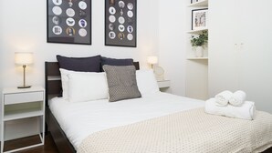Une chambre d’hôtel bien rangée, avec un lit, deux tables de chevet, une petite table, une étagère murale agrémentée d’une plante et deux tableaux encadrés accrochés au mur.