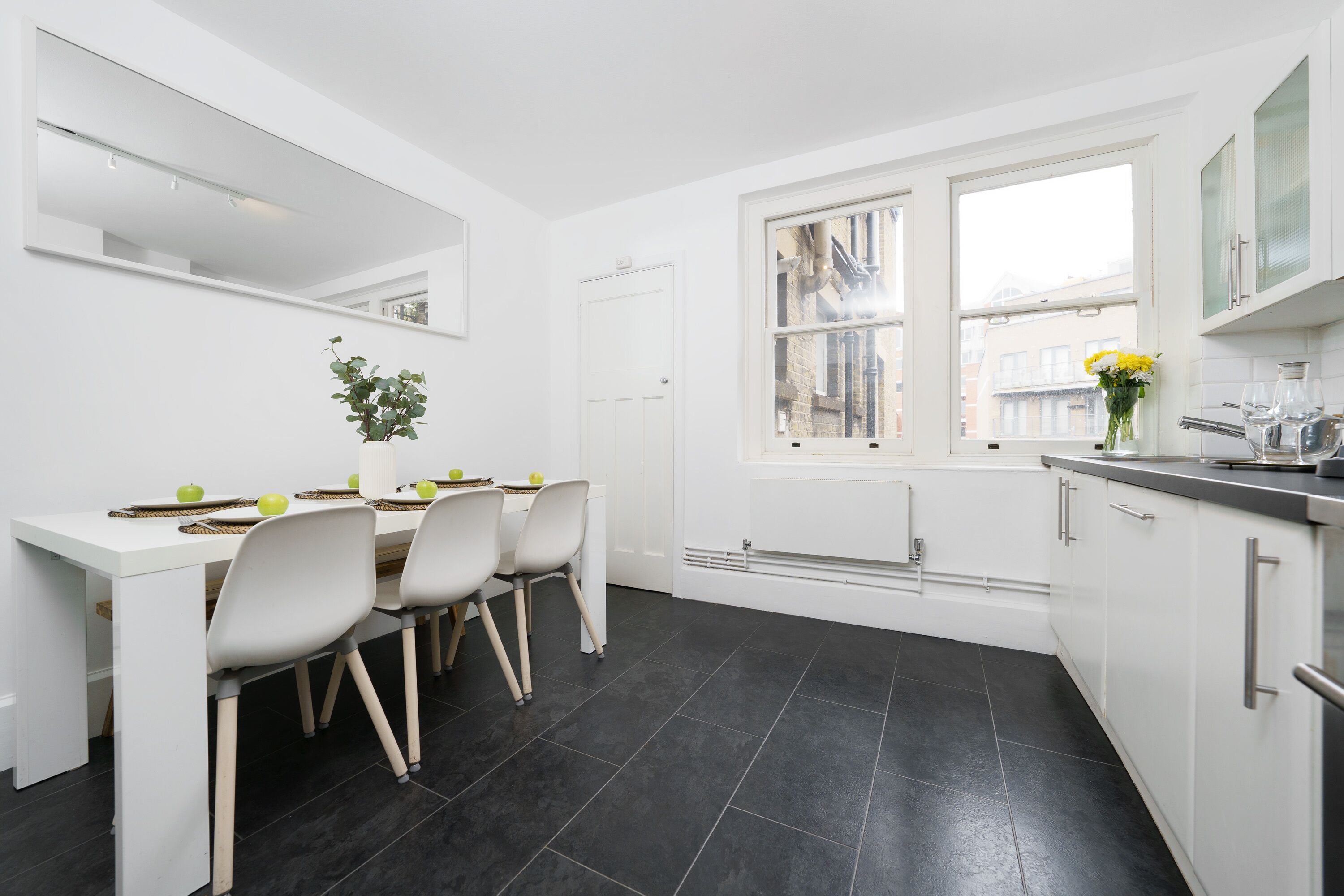 A modern kitchen with white cabinets, a dining table with chairs, a window with a view of buildings, and a plant on the table.