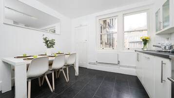 A modern kitchen with white cabinets, a dining table with chairs, a window with a view of buildings, and a plant on the table.