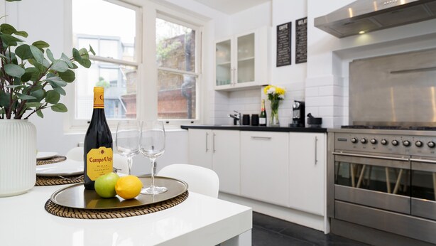 Une cuisine moderne avec des meubles blancs, un four en acier inoxydable et un coin repas agrémenté d’une table blanche, d’une bouteille de Campo Viejo, d’un verre et de deux citrons.