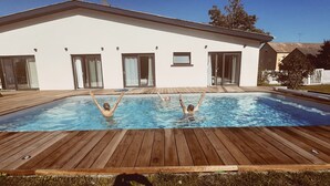 Uma piscina com duas pessoas, rodeada por um deque de madeira e um edifício branco com janelas amplas e portas de correr.