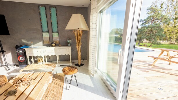 Um interior moderno com um banco de madeira, uma cômoda branca e um abajur. Uma porta de vidro dá acesso a uma área externa com piscina e um deck de madeira.