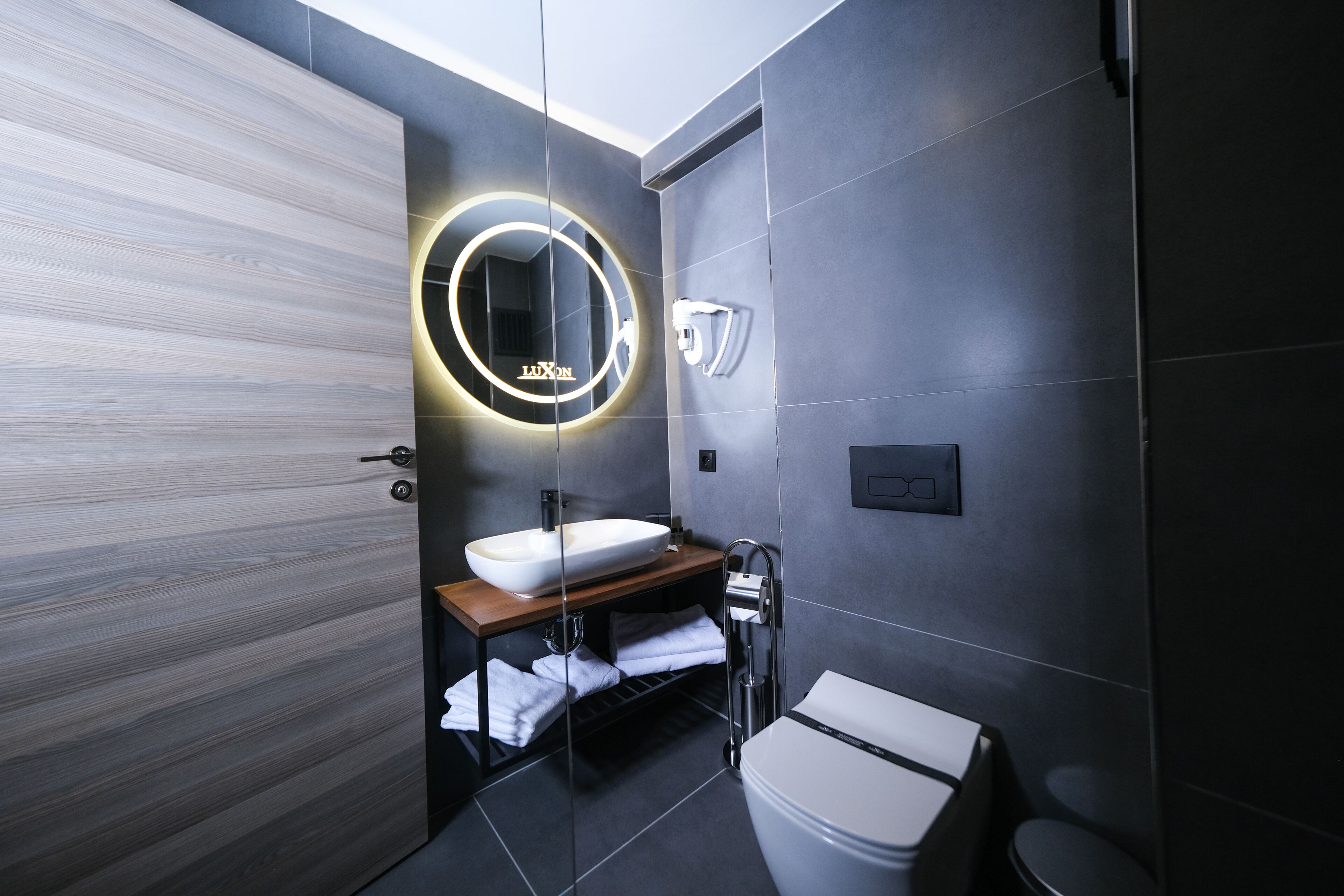 A modern bathroom with a wooden vanity, a round mirror with a light, a white sink, and a toilet.