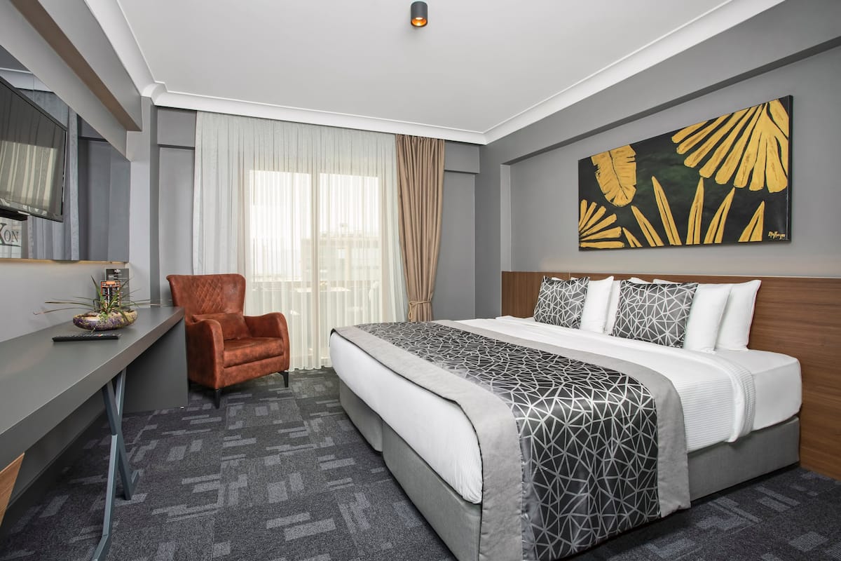 A hotel room with a bed, a desk, a chair, a large window with curtains, and a wall art piece.