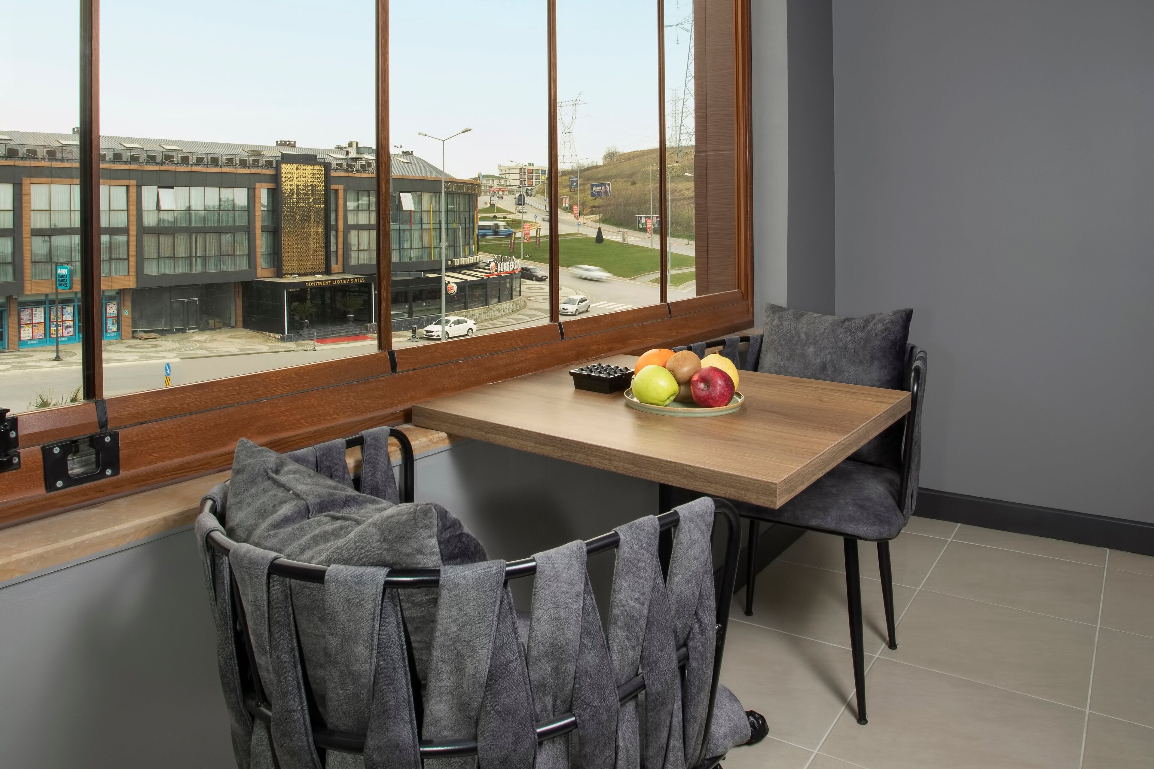 Une chambre d’hôtel moderne dotée d’une grande fenêtre donnant sur la rue et les immeubles. On y trouve une table en bois sur laquelle est posé un panier de fruits, ainsi qu’une chaise sur laquelle est drapée une serviette.