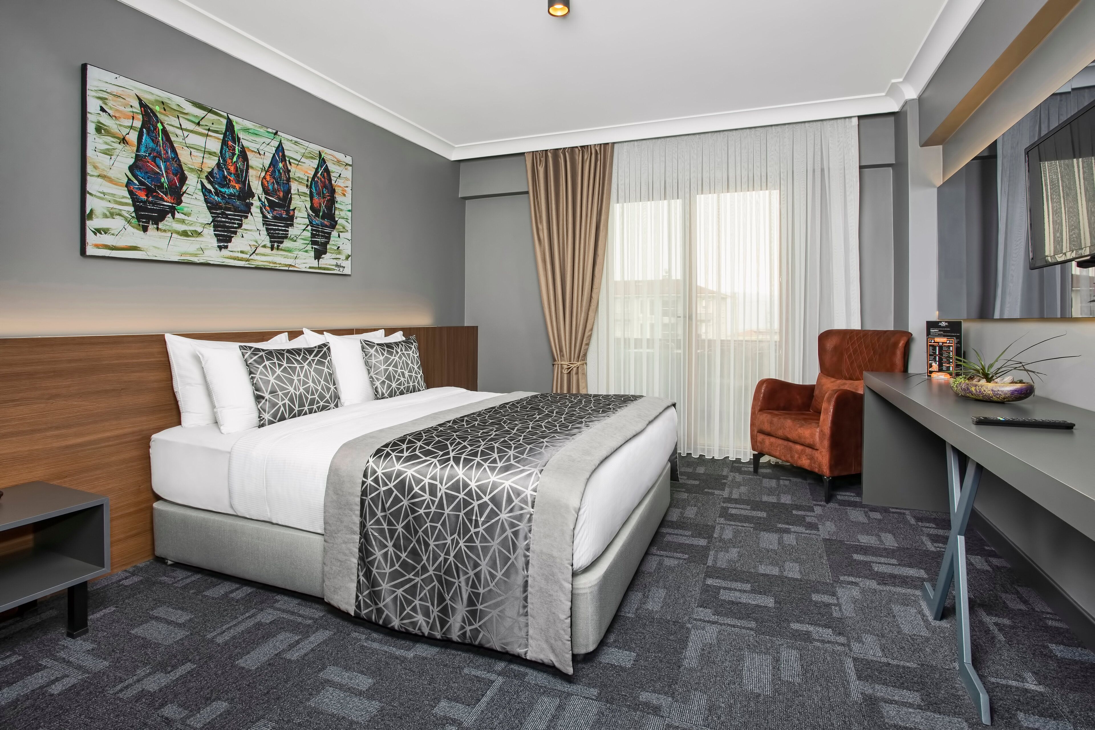 A modern hotel room with a large bed, a desk, a chair, and a painting on the wall.