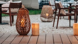 Een terras met houten tafels en stoelen, twee rieten lampen en een kaars.