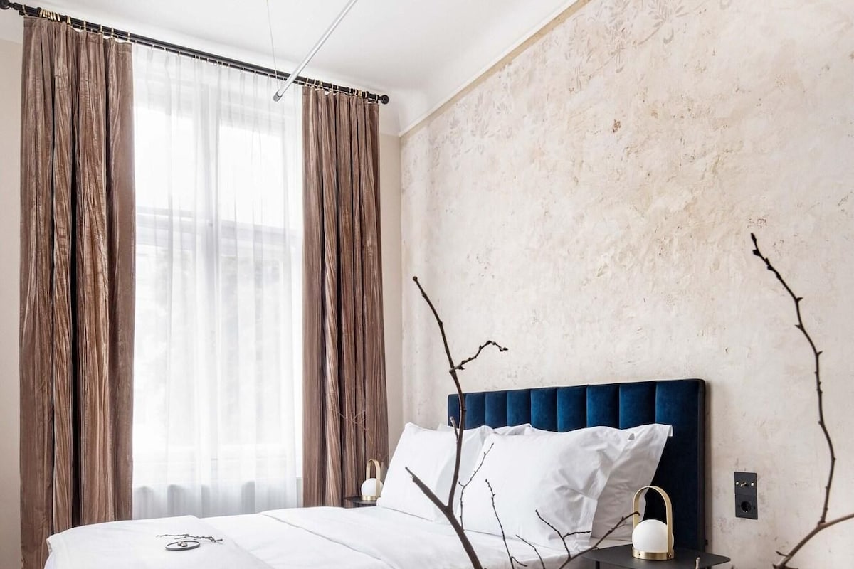 Une chambre d’hôtel moderne avec un grand lit, une table de chevet avec une lampe et une vue sur la ville à travers de fines rideaux.