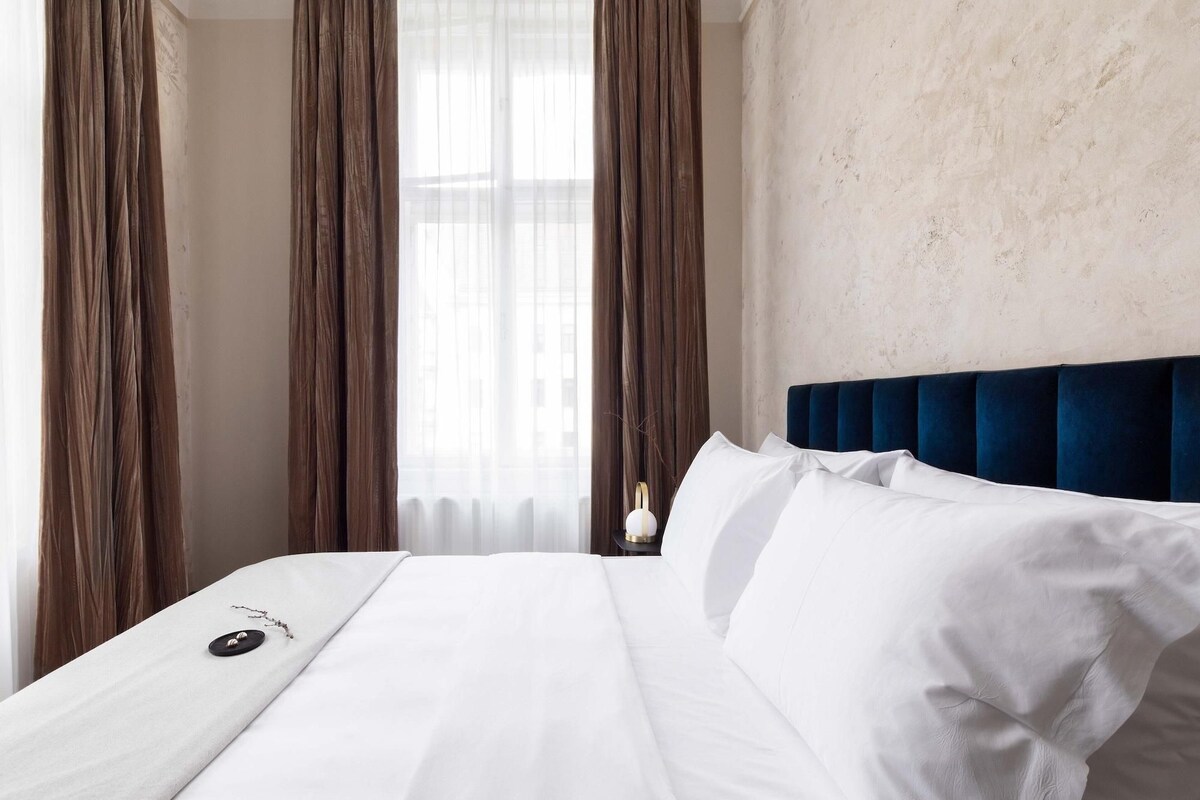 Een hotelkamer met een groot bed, witte lakens, een donkerblauw hoofdeinde en dunne gordijnen.