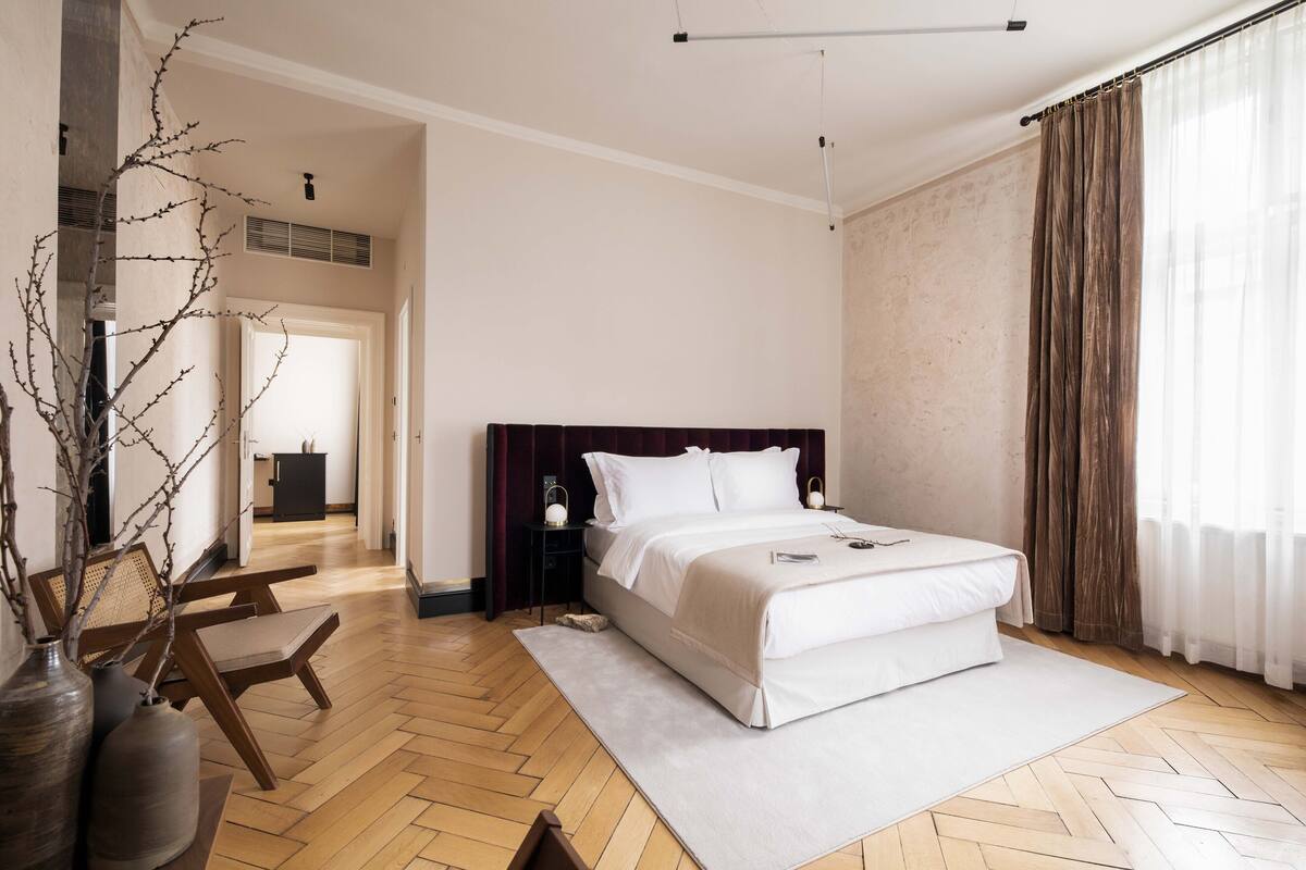 Une chambre avec un grand lit, un plancher en bois, une table de chevet, une lampe et une vue sur une autre pièce par une porte ouverte.
