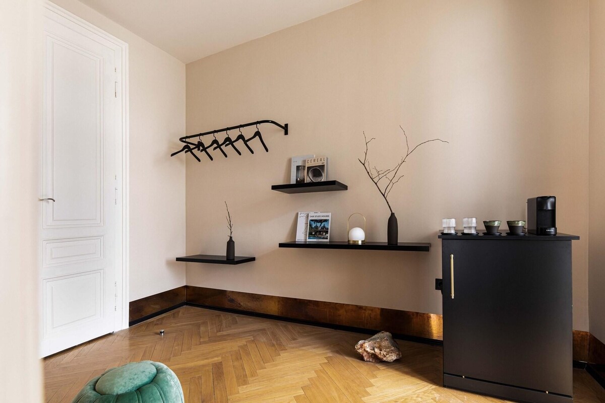 Een moderne keuken met een houten visgraatvloer, een zwarte koelkast en minimalistische inrichting, waaronder een zwart wandplankje met een boek en een kleine vaas.