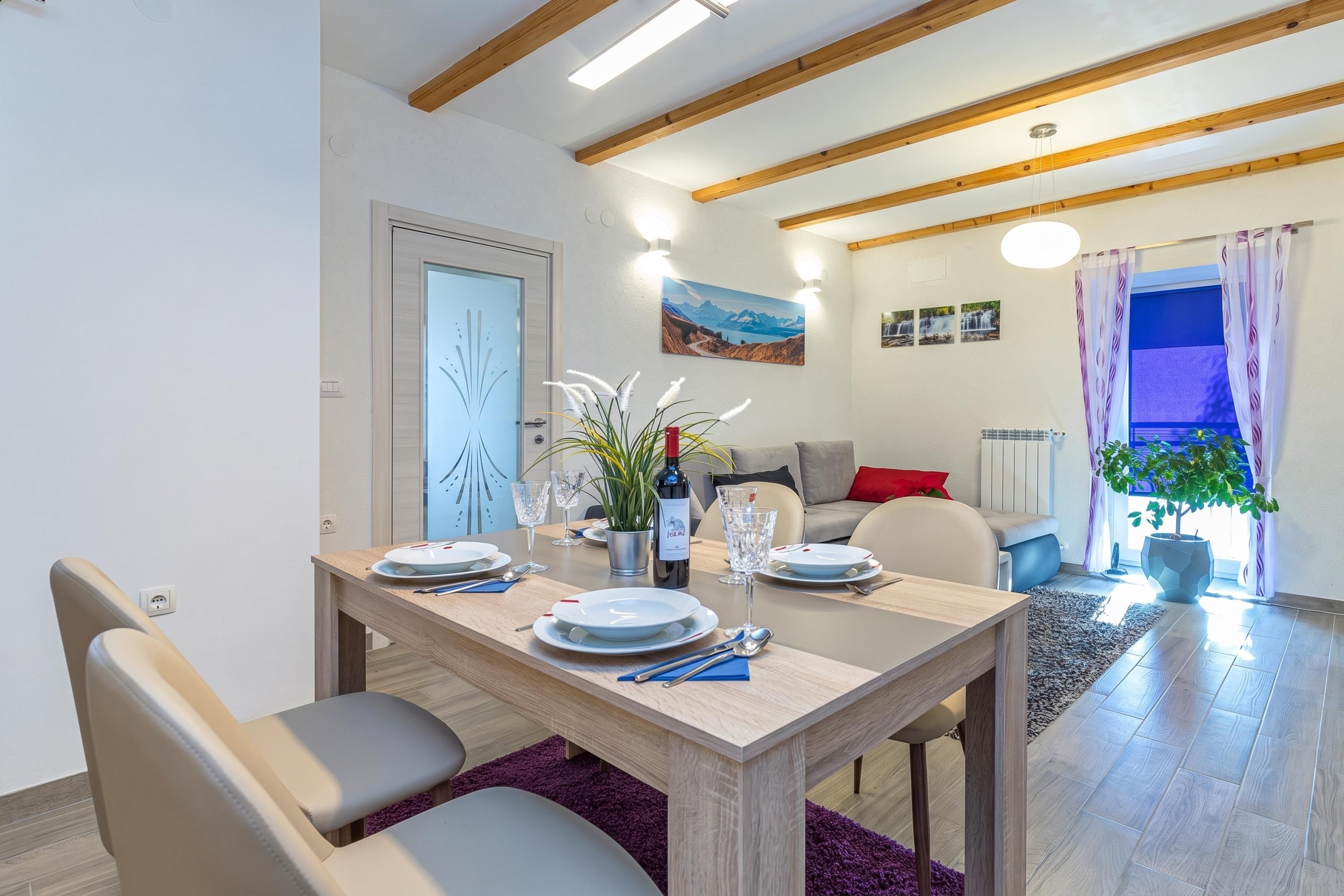 Un comedor con una mesa de madera dispuesta para cuatro personas, un sofá y un ventanal con cortinas.