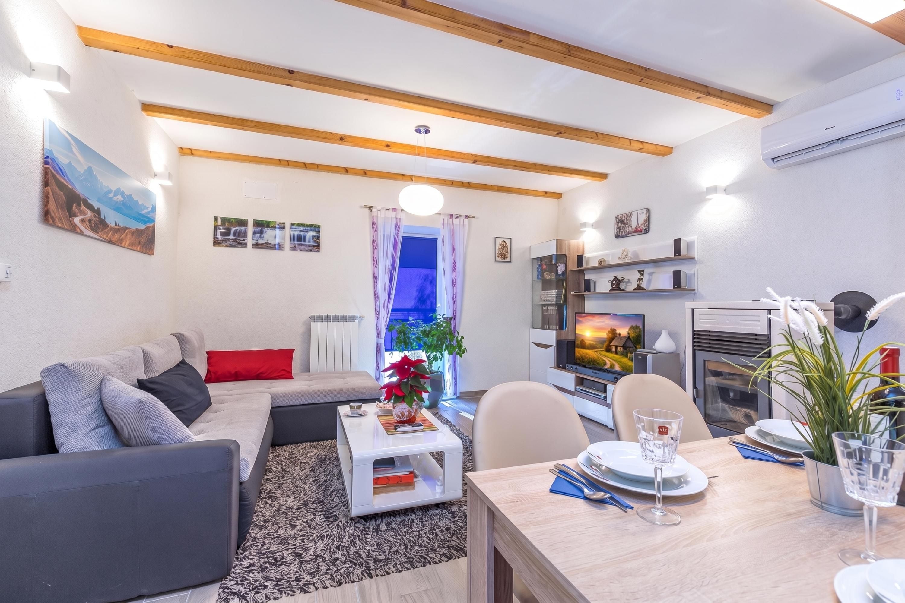 Una sala moderna con un sofá, una mesa de centro y un comedor. Hay un televisor, una chimenea y una ventana con cortinas.