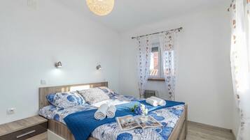 Una cama bien hecha con una colcha estampada, una mesita de noche con cajón, una lámpara de pared y una ventana con cortina.