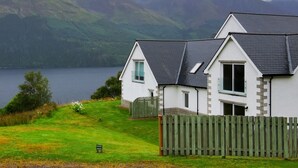 Une maison à deux étages, avec un toit sombre, des murs blancs et une clôture en bois à l’avant. Elle est située près d’une étendue d’eau, avec des montagnes en arrière-plan.