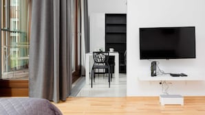 Una sala de estar moderna con un ventanal grande, un televisor de pantalla plana montado en la pared, una mesa blanca con dos sillas y un mueble blanco pequeño debajo del televisor.