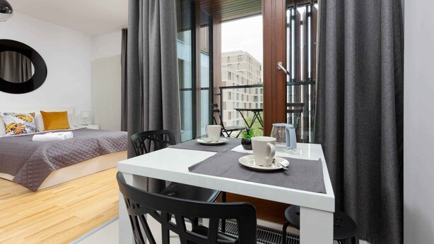 Una habitación de hotel moderna con una cama, una mesa de comedor con sillas, una cafetera y vistas a edificios a través de ventanas amplias.