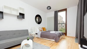 Un dormitorio moderno con un sofá gris, una mesita redonda blanca, una cama con colcha gris, un espejo redondo y un televisor sobre un mueble.