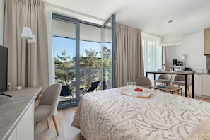 A modern hotel room with a large bed, a desk, a chair, a dining table, and a view of the outdoors.