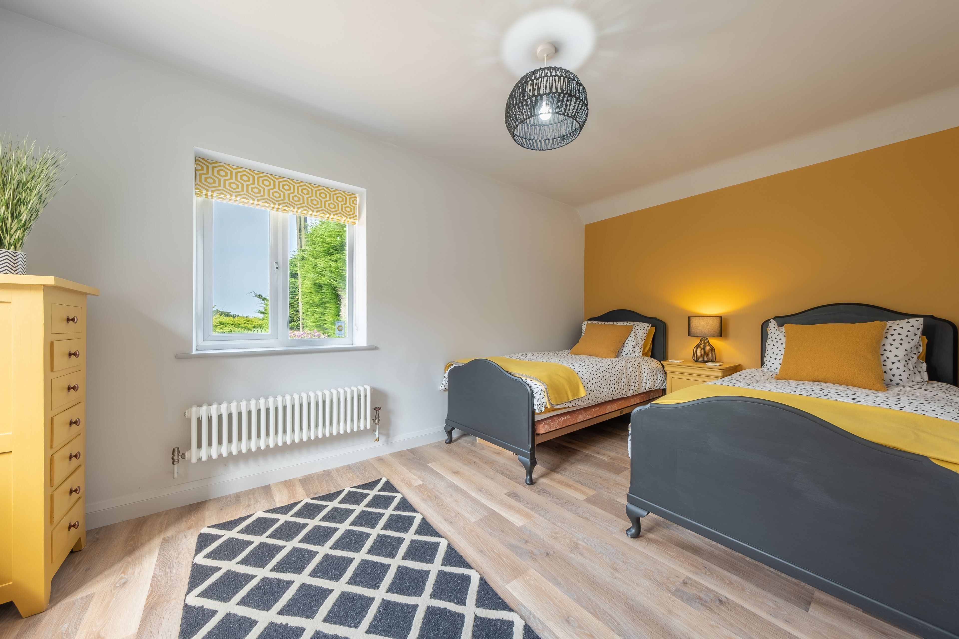 A bedroom with two beds, a radiator, a window with patterned curtains, a yellow dresser, and a geometric rug.