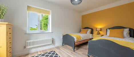 A bedroom with two beds, a radiator, a window with patterned curtains, a yellow dresser, and a geometric rug.