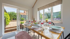 A dining area with a table set for four, large windows, and a view of the garden. - Castle Bungalow, Thornham, Norfolk (Thornham)