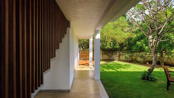 Un corredor exterior cubierto con una pared de listones de madera en un lado, que conduce a un área con césped, un árbol y un banco.