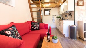 A compact living space with a red sofa, a wooden table with a bottle and glass, and a kitchen area with cabinets and a trash bin. - Modern family&pet friendly villa with private sauna and beach (Bosljiva Loka)